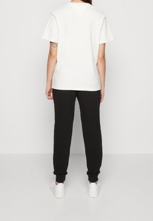 Henkilö pukeutunut yksiväriseen valkoiseen lyhythihaiseen t-paitaan, mustiin housuihin, valkoiset tennarit jalassa, seisoo selin päin näyttäen hämähäkinverkkotatuoinnin vasemmassa kädessä.