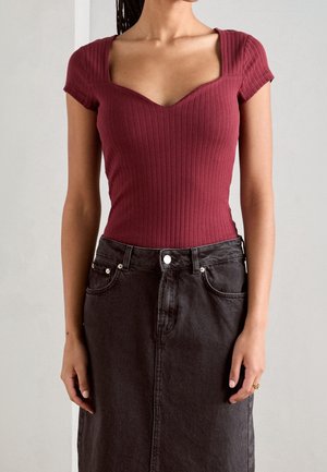 Vrouw draagt een nauwsluitend geribd bordeaux topje met korte mouwen en een donkere high-waisted denim rok, staand tegen een effen achtergrond.