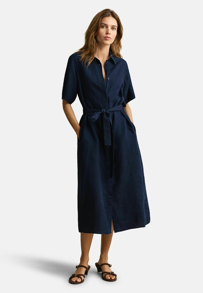 Femme portant une robe midi bleu marine boutonnée avec manches courtes et ceinture à la taille, debout les mains dans les poches, associée à des sandales à talons marron.