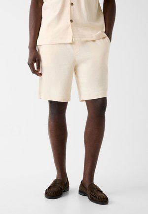 Mann trägt cremefarbene Shorts, passendes Hemd mit Knöpfen und dunkelbraune Slip-on-Loafers vor einfachem Hintergrund.