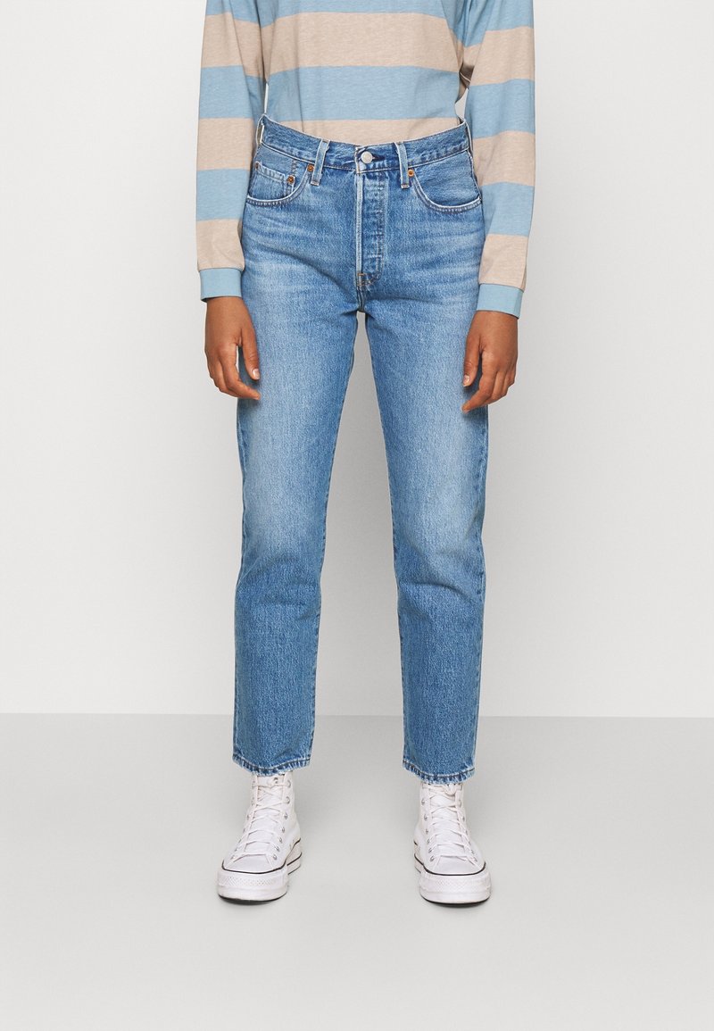 Persona vistiendo una camisa de manga larga a rayas en azul claro y beige, jeans de tiro alto en azul y zapatillas blancas de caña alta contra un fondo liso.