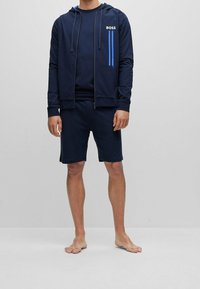 Uomo che indossa un felpa blu navy con il testo "BOSS" e pantaloni corti blu navy abbinati, in piedi a piedi nudi contro uno sfondo grigio semplice.