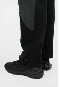Czarne spodnie sportowe z szarym panelem, z troczkiem przy kostce. Noszone z czarnymi sneakersami o fakturowanej powierzchni i wyraźnych podeszwach.