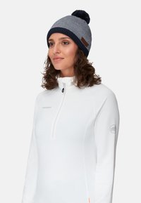 Marinblå och grå texturerad stickad mössa med en pom-pom, med en läderlogotyp-patch. Bärs med en vit zip-up tröja.