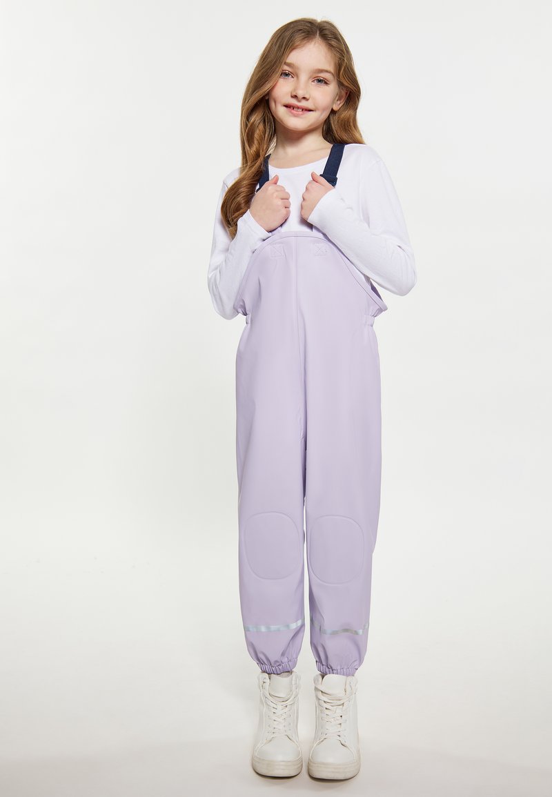 Jauna mergaitė su ilgais plaukais, vilkinti violetinius vandeniui atsparius kombinezonus, baltą ilgų rankovių marškinį ir baltus batus, laiko kombinezono diržus ir šypsosi.