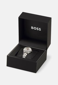 Sølv rustfritt stål klokk med teksturert skive og lenkearmbånd, vist i en svart boks med sølv "BOSS" logo.