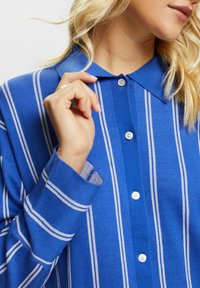 Camisa de botones a rayas azules con un cuello clásico, rayas blancas y botones en la parte delantera. Textura suave con un corte relajado.