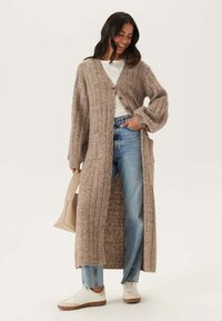 Lange, ribbelige cardigan in lichtbruin, gemaakt van zachte stof. Heeft knopen aan de voorkant, twee zakken en brede mouwen. Draagt over een witte top.