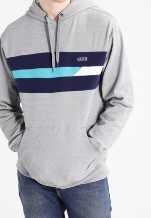 Mann trägt grauen Vans Hoodie mit navyblauen, türkisen und weißen Horizontalstreifen, blaue Jeans, Hände in der vorderen Tasche.