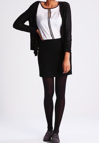 Frau in einem schwarzen Cardigan über einer weißen Bluse mit schwarzem Rand, schwarzer Rock, schwarze Strumpfhose und dunklen Slippern, steht mit einer Hand in der Hüfte.