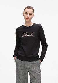 Femme portant un sweat noir avec le logo "Karl" et un pantalon à carreaux gris, se tenant avec les mains dans les poches devant un fond blanc.
