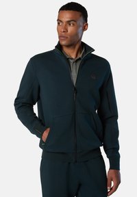 Sudadera con cremallera de color verde oscuro, hecha de tejido liso, que cuenta con un cuello alzado, dos bolsillos frontales y un pequeño logo en el pecho.
