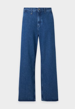 Joven con chaqueta de cuero negra, camiseta blanca, vaqueros azules anchos y zapatos negros, de pie con las manos en los bolsillos.