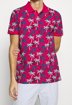 Man wearing bright pink polo shirt with white and blue palm tree pattern and white shorts, standing against a plain background.