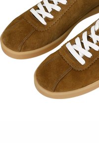 Braune Wildledersneaker mit weißen Schnürsenkeln, umrandeter Naht, runder Zehenbereich und einer hellbraunen Gummisohle. Texturierte Oberfläche und simples Design.