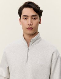 Gaiši pelēks džemperis, kas izgatavots no mīksta auduma ar rāvējslēdzēja apkakli. Iekļauj izšūtu logo uz krūtīm un brīvu piegriezumu ar nolaižamām plecu šuvēm.