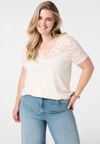 Korte mouwen crème top met kant inzet en een v-hals, gecombineerd met lichtblauwe spijkerbroek. Soepele textuur met een relaxte pasvorm.