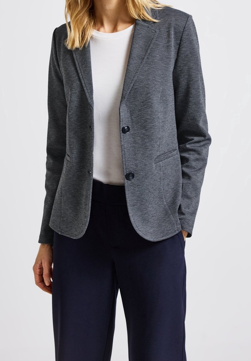 Femme portant un blazer gris cintré sur une chemise blanche et un pantalon bleu marine foncé, debout avec une main dans la poche.