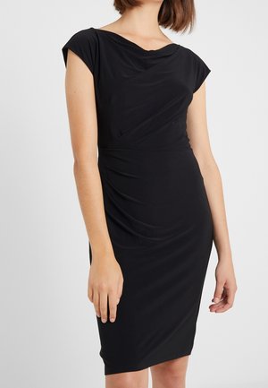 Woman wearing a fitted black sleeveless dress with draped neckline and gathered details on the waist, standing against a plain background.