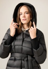 Czarna pikowana kurtka puchowa z wcięciem w talii, wyposażona w kaptur i czarne akcenty. Gładka, pikowana tekstura z paskiem w talii.