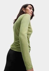 Maglietta aderente a maniche lunghe in tessuto verde chiaro, dalla superficie liscia. La modella la indossa con una gonna nera, vista di profilo su sfondo bianco.