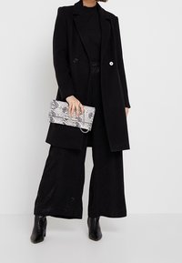 Manteau noir long avec deux boutons, associé à un pantalon large texturé noir et des bottines. Tenant un sac à main en cuir python.
