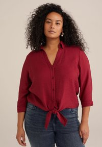Camicia rossa con bottoni, dettaglio con nodo anteriore, realizzata in tessuto strutturato, con scollo a V e maniche a tre quarti. Abbinata a un paio di jeans.