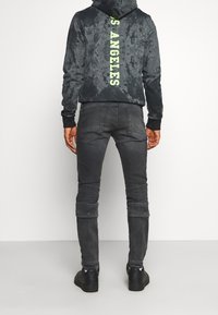Person med svarta Nike-skor, mörkgrå jeans med knäflikdetaljer och en svart hoodie med "S. ANGELES" i gul text på baksidan.