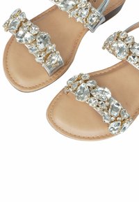 Sandali in pelle beige con un cinturino prominente decorato da cristalli, composto da forme a goccia e rotonde. Dettagli in metallo argentato visibili.