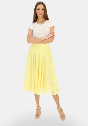 Kobieta stojąca ze skrzyżowanymi ramionami, ubrana w biały krótki rękaw, żółtą spódnicę midi i beżowe szpilki, na jednolitym tle.