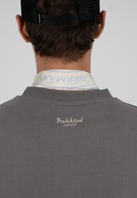 Homme portant une casquette noire et une chemise grise avec le mot « Interdit » brodé dans le dos, et un col blanc avec un texte bleu clair visible au-dessus de l'encolure de la chemise.