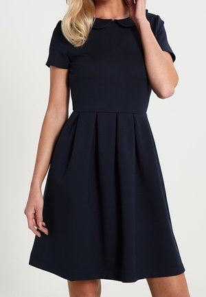 Frau trägt ein marineblaues, knielanges Kleid mit kurzen Ärmeln, Falten und einem dezenten Kragen.