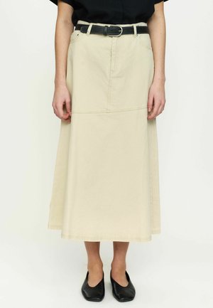 Woman wearing a beige midi skirt with front seam and black belt, paired with a black top and black flat shoes, standing.