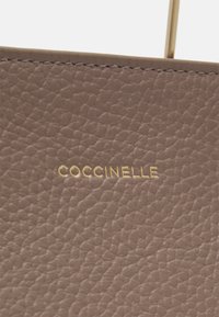 Borsa in pelle color taupe con superficie testurizzata. Presenta il logo dorato "COCCINELLE", che mette in evidenza un design minimalista e linee pulite.