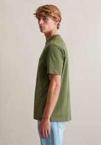 T-shirt bawełniany w oliwkowo-zielonym kolorze z krótkimi rękawami i okrągłym dekoltem, zestawiony z jasnoniebieskimi dżinsami, widoczny z profilu.