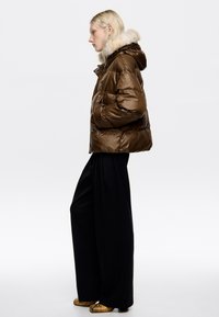 Hnedá nafukovacia bunda s lesklým povrchom a kapucňou s kožušinkou, skombinovaná s čiernymi nohavicami s širokými nohami a obuvou s zlatými akcentmi.