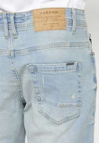 Jean en denim bleu clair avec une finition texturée, présentant une étiquette en cuir à la taille et une légère usure sur les poches et les coutures.
