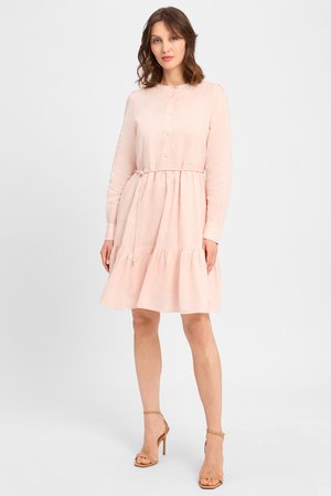 Frau trägt ein hellrosa, langärmliges, geknöpftes Kleid mit Taillengürtel und gerafftem Saum, kombiniert mit nudefarbenen Sandalen mit hohen Absätzen, stehend.