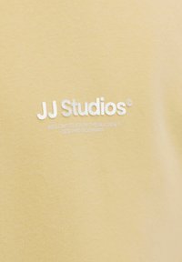 Tissu en coton jaune à texture lisse, avec un texte blanc indiquant « JJ Studios » et d'autres détails de marque en dessous.