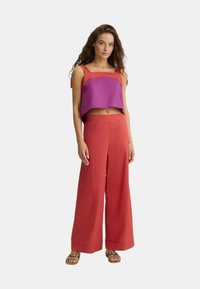 Top corto viola e rosso con spalline spesse, abbinato a pantaloni rossi a gamba larga e sandali marroni con tacchetti decorati.