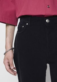 Manšestrové kalhoty tmavě námořnické barvy s žebrovanou texturou, stříbrné knoflíkové zdobení a boční kapsa. Ruka ukazuje kovový náramek.