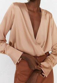 Satinbluse i blød beige med dyb V-hals, lange ærmer og knaplukninger ved håndleddene, parret med højtaljede brune bukser.