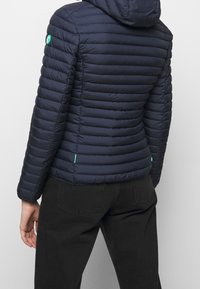 Henkilö, jolla on yllään istuva, vuorattu tummansininen huppujakku ja mustat farkut, näkyy selästä yksinkertaista vaaleaa taustaa vasten.