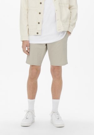 Shorts beige clair en tissu lisse et léger, avec une coupe droite et des poches latérales, portés avec un t-shirt blanc à col rond et une veste.