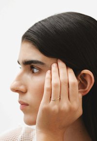 Anillo de oro con una pequeña piedra azul, usado en el dedo anular, contra un perfil en primer plano de una persona con cabello oscuro y un fondo neutro.