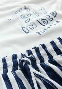 T-shirt de algodón blanca con el texto "Quedémonos en la cama para siempre" en azul, combinada con pantalones cortos de pijama a rayas en navy y blanco con cinturilla elástica.