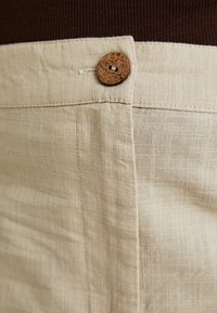 Pantalones de lino beige con una superficie texturizada, que cuentan con un botón de corcho en la cintura y un sutil diseño tejido en la tela.