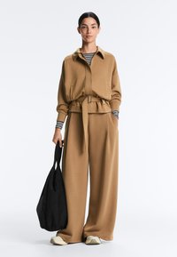 Kvinde iført kamelfarvet jakke med bælte og bukser med vide ben, stribet langærmet top, hvide sneakers, holder en sort taske i højre hånd.