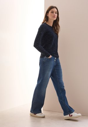 Chemise texturée à manches longues bleu marine associée à un jean bleu à large jambes, avec de grands boutons et des baskets blanches avec des accents rouges.