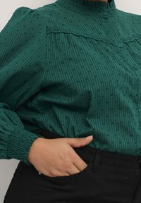 Groene textuur blouse met verticale strepen en kleine stippen, voorzien van een gerimpelde kraag en knoopsluiting. De mouwen zijn aan de polsen verzameld.